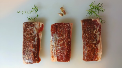 Three raw steaks seasoned with pepper are laid side by side on a light surface. Fresh herbs and pieces of garlic are placed artistically around the steaks for decoration.