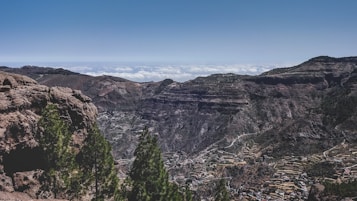 A panoramic view of a rugged mountain landscape with valleys and terraces. Pine trees appear in the foreground, while a backdrop of jagged peaks stretches into the horizon under a clear blue sky. Clouds are visible in the distance, hovering above the mountain range.