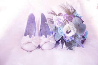 A pair of glittery white shoes adorned with large bows lies next to a vibrant bouquet of blue and purple flowers. The shoes have a delicate, feminine appearance, and they are placed on a soft, light-colored fabric. The bouquet includes a mix of roses and greenery, adding to the romantic and elegant atmosphere.
