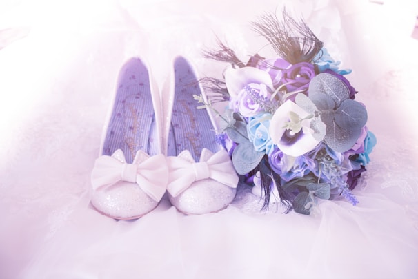 A pair of glittery white shoes adorned with large bows lies next to a vibrant bouquet of blue and purple flowers. The shoes have a delicate, feminine appearance, and they are placed on a soft, light-colored fabric. The bouquet includes a mix of roses and greenery, adding to the romantic and elegant atmosphere.