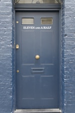 A dark blue door with the label 'ELEVEN AND A HALF' written in white above a mail slot. The door has two small windows near the top and a decorative semicircular window above. The surrounding wall is made of brick and painted the same shade of blue.