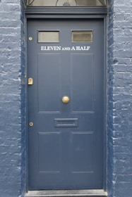 A dark blue door with the label 'ELEVEN AND A HALF' written in white above a mail slot. The door has two small windows near the top and a decorative semicircular window above. The surrounding wall is made of brick and painted the same shade of blue.