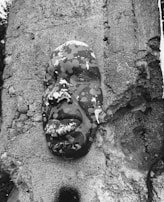 A sculpted face is embedded in a rough concrete wall. The mask-like feature is adorned with a camouflage pattern of light and dark spots, creating an intriguing blend with the surroundings. The material on the face appears weathered, suggesting exposure to the elements.