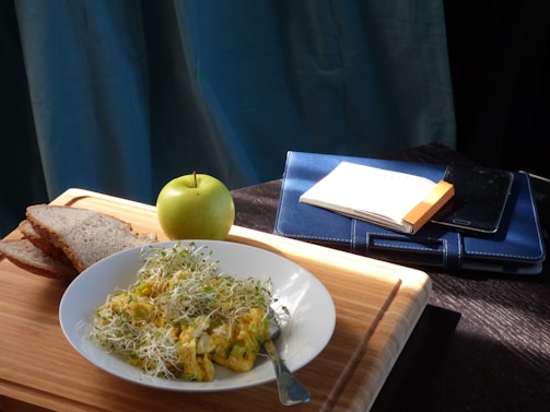 On a wooden surface, there is a plate of scrambled eggs topped with alfalfa sprouts, accompanied by slices of bread. Beside the plate, a green apple rests near a leather-covered notebook with a pen placed on top. A smartphone is partially visible under the notebook.