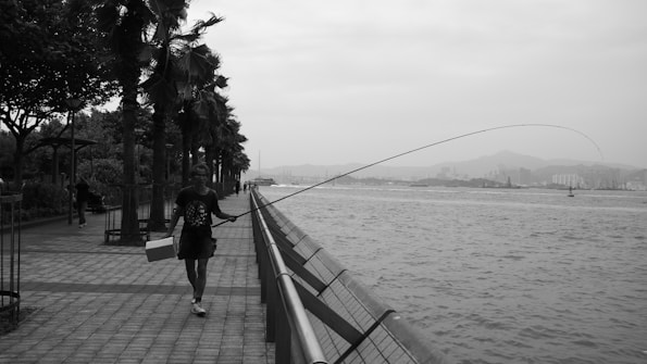 A black and white photograph of a person walking along a paved waterfront with a fishing pole in one hand and a box in the other. Palm trees line the pathway on the left, creating a serene and natural ambiance. The water stretches out to the right, and city buildings and mountains are visible in the hazy background.