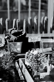 A monochrome image of a market stall display featuring small floral bouquets labeled as 'Spring Posies' with a price tag of £2.00. The flowers are arranged in a transparent glass bottle. Behind the display, there are additional containers, possibly with more plants or flowers, and a series of vertical wooden structures in the background.