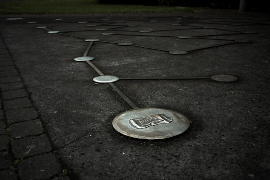 Metallic discs connected by lines are embedded in a dark, paved surface, forming a geometric pattern. The ground appears slightly wet, and there is greenery visible at the edge. One disc prominently features an engraved image of a can.
