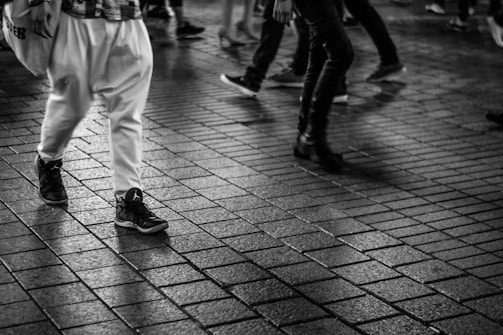 A black and white image featuring multiple people walking on a tiled street surface. Focus is on the lower legs and feet, showcasing a variety of footwear styles including sneakers and boots. The ground appears wet, reflecting parts of the figures.