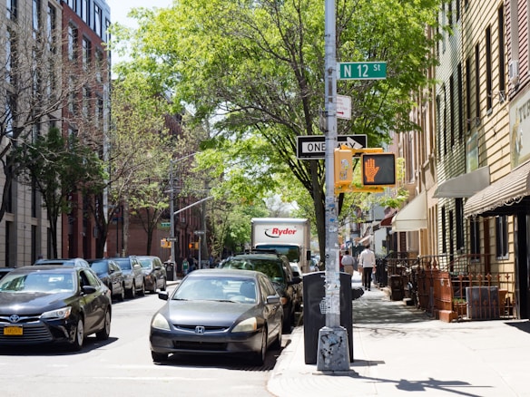 A street scene featuring a row of parked cars along both sides. There is a traffic signal and street signs, including one indicating N 12 St and a 'one way' sign. People can be seen walking on the sidewalk. Trees with green foliage line the street, and various buildings, including residential and commercial properties, are visible along the sidewalk. A delivery truck with the Ryder logo is also present in the street.