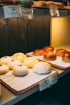 A display of assorted bread rolls arranged neatly on trays, featuring a variety of textures and toppings. The lighting is warm and gives the bakery a cozy atmosphere, with labels in Japanese above each type of bread.