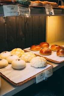 A display of assorted bread rolls arranged neatly on trays, featuring a variety of textures and toppings. The lighting is warm and gives the bakery a cozy atmosphere, with labels in Japanese above each type of bread.