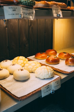A display of assorted bread rolls arranged neatly on trays, featuring a variety of textures and toppings. The lighting is warm and gives the bakery a cozy atmosphere, with labels in Japanese above each type of bread.
