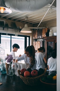 Several people are gathered in a cozy kitchen space, engaged in preparing food or drinks. The kitchen has a warm atmosphere with a wooden table, a basket of fruit, floral arrangements, and various kitchen utensils. Natural light flows in through large windows, and overhead lamps provide additional lighting.