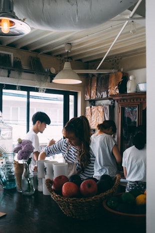Several people are gathered in a cozy kitchen space, engaged in preparing food or drinks. The kitchen has a warm atmosphere with a wooden table, a basket of fruit, floral arrangements, and various kitchen utensils. Natural light flows in through large windows, and overhead lamps provide additional lighting.
