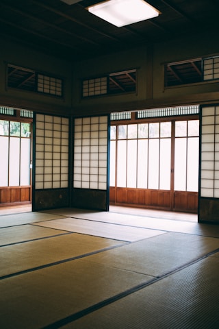 A traditional Japanese room with tatami mats covering the floor and shoji screens allowing natural light to filter in. The room has wooden elements and clean lines, creating a serene and minimalist environment.