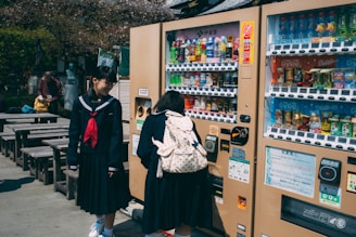 Two students in school uniforms are standing near vending machines. One is selecting a drink while the other is watching with a smile. The setting includes outdoor wooden benches and some trees in the background.