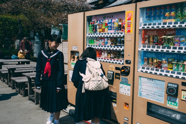 Two students in school uniforms are standing near vending machines. One is selecting a drink while the other is watching with a smile. The setting includes outdoor wooden benches and some trees in the background.