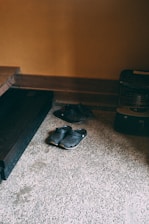 A pair of black clogs placed on a speckled stone floor near a corner of a room with warm, light brown walls. Near the footwear is a dark wooden shelf and a small black heater.