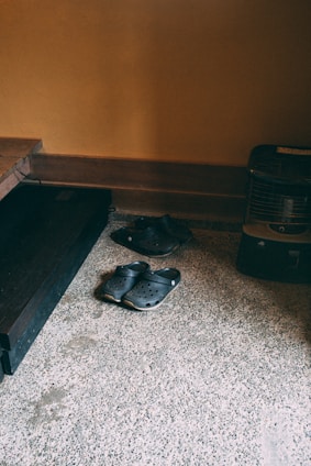 A pair of black clogs placed on a speckled stone floor near a corner of a room with warm, light brown walls. Near the footwear is a dark wooden shelf and a small black heater.
