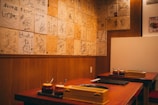 A cozy restaurant interior featuring wooden walls adorned with numerous signed plaques or pieces of paper. Two tables are visible with sauce containers and cooking equipment, suggesting a setting for communal dining or a barbecue style restaurant.