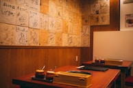 A cozy restaurant interior featuring wooden walls adorned with numerous signed plaques or pieces of paper. Two tables are visible with sauce containers and cooking equipment, suggesting a setting for communal dining or a barbecue style restaurant.