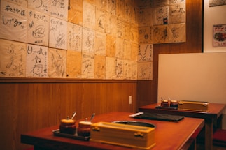 A cozy restaurant interior featuring wooden walls adorned with numerous signed plaques or pieces of paper. Two tables are visible with sauce containers and cooking equipment, suggesting a setting for communal dining or a barbecue style restaurant.
