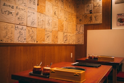 A cozy restaurant interior featuring wooden walls adorned with numerous signed plaques or pieces of paper. Two tables are visible with sauce containers and cooking equipment, suggesting a setting for communal dining or a barbecue style restaurant.