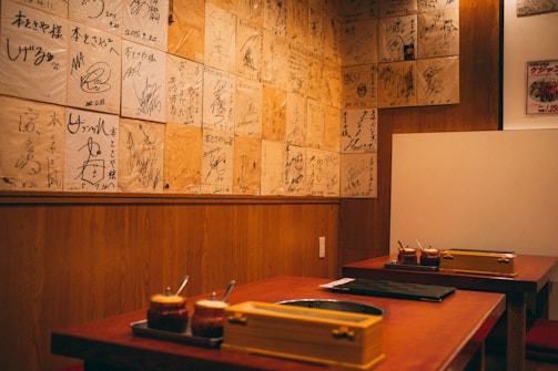 A cozy restaurant interior featuring wooden walls adorned with numerous signed plaques or pieces of paper. Two tables are visible with sauce containers and cooking equipment, suggesting a setting for communal dining or a barbecue style restaurant.