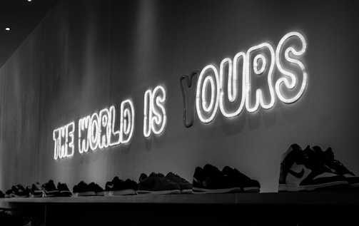 A series of sneakers are aligned on a shelf beneath a neon sign that reads 'THE WORLD IS YOURS'. The scene is captured in black and white, giving a modern and edgy feel.