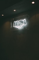 A neon sign depicting a handshake is mounted on a dark wall, illuminated by overhead lights.