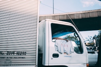 A white truck is parked with a t-shirt hanging from the side mirror, displaying graphic designs. The truck is positioned under an overpass, with clear blue skies visible above. The side panel of the truck features text and numbers.