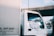 A white truck is parked with a t-shirt hanging from the side mirror, displaying graphic designs. The truck is positioned under an overpass, with clear blue skies visible above. The side panel of the truck features text and numbers.
