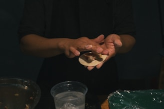 A person is carefully shaping dough filled with what appears to be a savory mixture, possibly preparing dumplings or similar food. The hands are slightly dusted with flour, and there is a bowl with more fillings and a container possibly with water. The lighting is dim, focusing primarily on the hands and the dough.