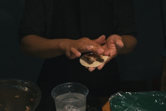 A person is carefully shaping dough filled with what appears to be a savory mixture, possibly preparing dumplings or similar food. The hands are slightly dusted with flour, and there is a bowl with more fillings and a container possibly with water. The lighting is dim, focusing primarily on the hands and the dough.