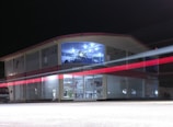 A two-story building with glass-paneled walls is illuminated in the evening, exhibiting an interior filled with shelves and various products. A blurred red light from a passing vehicle creates a streak across the front, adding a dynamic element against the stillness of the structure. The upper section has a distinctive logo and is well-lit, suggesting a commercial or business environment.