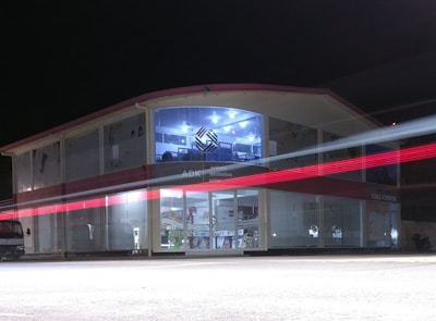 A two-story building with glass-paneled walls is illuminated in the evening, exhibiting an interior filled with shelves and various products. A blurred red light from a passing vehicle creates a streak across the front, adding a dynamic element against the stillness of the structure. The upper section has a distinctive logo and is well-lit, suggesting a commercial or business environment.