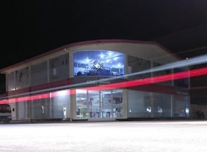 A two-story building with glass-paneled walls is illuminated in the evening, exhibiting an interior filled with shelves and various products. A blurred red light from a passing vehicle creates a streak across the front, adding a dynamic element against the stillness of the structure. The upper section has a distinctive logo and is well-lit, suggesting a commercial or business environment.