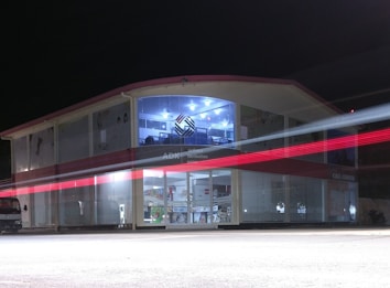 A two-story building with glass-paneled walls is illuminated in the evening, exhibiting an interior filled with shelves and various products. A blurred red light from a passing vehicle creates a streak across the front, adding a dynamic element against the stillness of the structure. The upper section has a distinctive logo and is well-lit, suggesting a commercial or business environment.