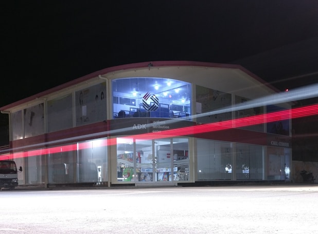 A two-story building with glass-paneled walls is illuminated in the evening, exhibiting an interior filled with shelves and various products. A blurred red light from a passing vehicle creates a streak across the front, adding a dynamic element against the stillness of the structure. The upper section has a distinctive logo and is well-lit, suggesting a commercial or business environment.