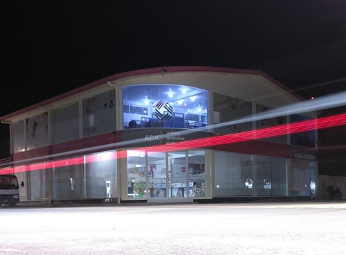 A two-story building with glass-paneled walls is illuminated in the evening, exhibiting an interior filled with shelves and various products. A blurred red light from a passing vehicle creates a streak across the front, adding a dynamic element against the stillness of the structure. The upper section has a distinctive logo and is well-lit, suggesting a commercial or business environment.