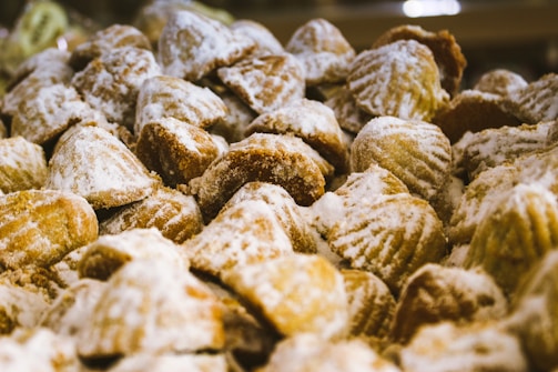 A close-up view of a large pile of pastries covered in powdered sugar. The pastries have a shell-like shape and appear to be freshly baked, with a golden brown color visible beneath the sugar coating.