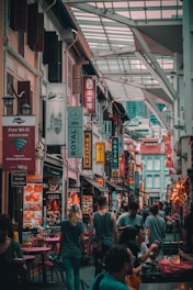 A bustling street in a market with numerous signs in multiple languages. People are walking and seated at tables, suggesting a lively urban atmosphere. The architecture features a mix of traditional and modern styles, with vibrant colors and decorations.