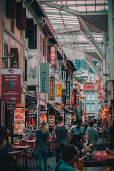 A bustling street in a market with numerous signs in multiple languages. People are walking and seated at tables, suggesting a lively urban atmosphere. The architecture features a mix of traditional and modern styles, with vibrant colors and decorations.