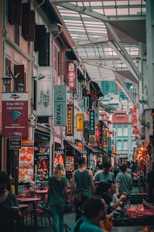 A bustling street in a market with numerous signs in multiple languages. People are walking and seated at tables, suggesting a lively urban atmosphere. The architecture features a mix of traditional and modern styles, with vibrant colors and decorations.