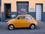 A classic, small yellow car is parked on a street in front of a building wall covered with colorful graffiti. The building has a distinct urban look with its grey lower half and yellow upper half, featuring roll-up doors and a glass entrance door.
