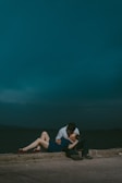 A couple sharing an intimate moment against a moody, dark blue sky. The woman is lying down with her legs extended, wearing a blue dress and red shoes, while the man leans over her wearing a shirt and dark pants.