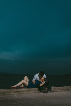 A couple sharing an intimate moment against a moody, dark blue sky. The woman is lying down with her legs extended, wearing a blue dress and red shoes, while the man leans over her wearing a shirt and dark pants.