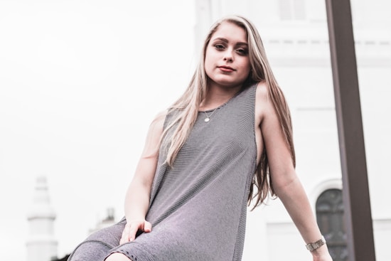 A young woman with long hair is looking confidently at the camera, wearing a sleeveless dress. She is positioned outdoors against a light, blurred architectural background.