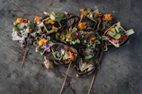Several traditional offerings are arranged on the ground, featuring vibrant flowers and leaves carefully arranged in small baskets made of natural materials. The background is a textured gray surface.