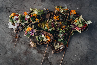 Several traditional offerings are arranged on the ground, featuring vibrant flowers and leaves carefully arranged in small baskets made of natural materials. The background is a textured gray surface.